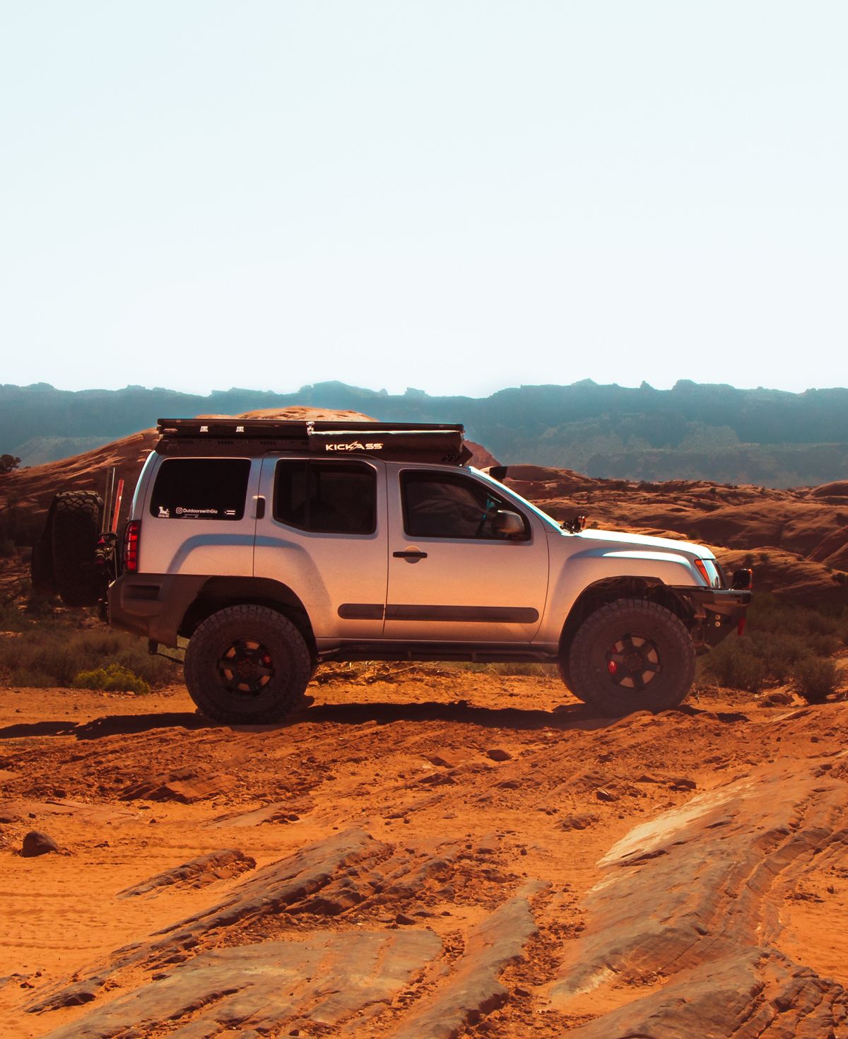 Silver Nissan Xterra with KickAss rooftop tent platform on Moab red rock at sunset