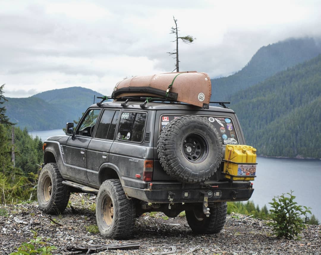 1981 Toyota Land Cruiser BJ60 in dark grey with a canoe strapped to the roof rack above a mountain lake