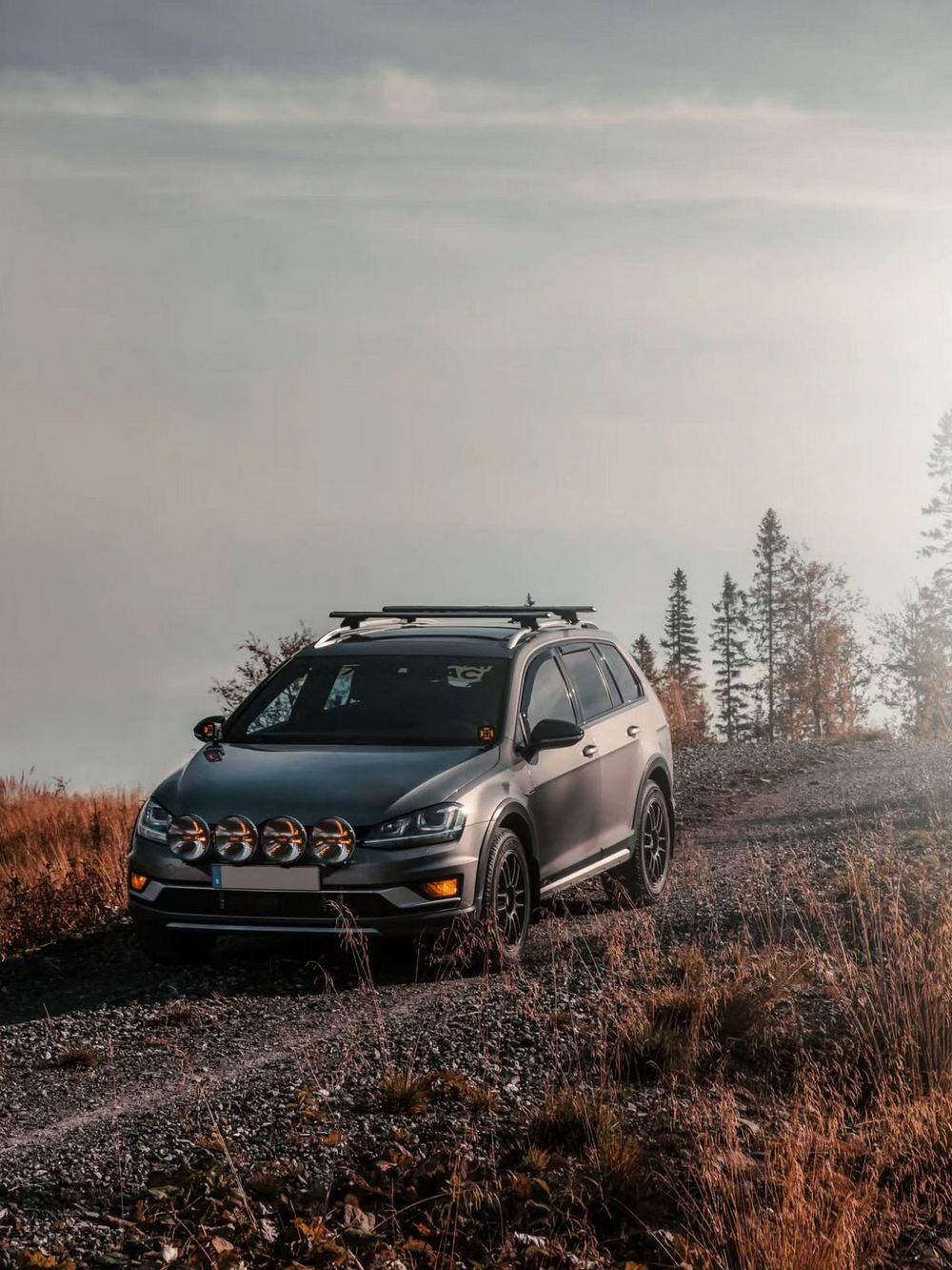 Lifted VW Golf Alltrack wagon with roof rack and rally lights positioned on gravel trail during overcast conditions