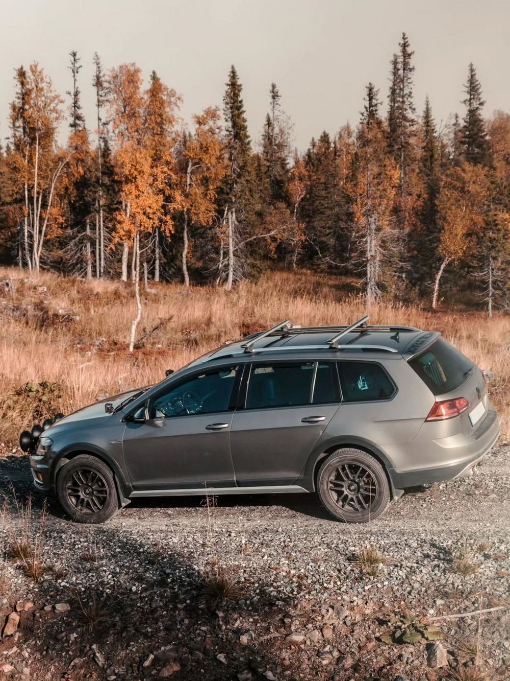 Lifted VW Golf Alltrack wagon with roof rack parked on gravel road beside autumn forest landscape